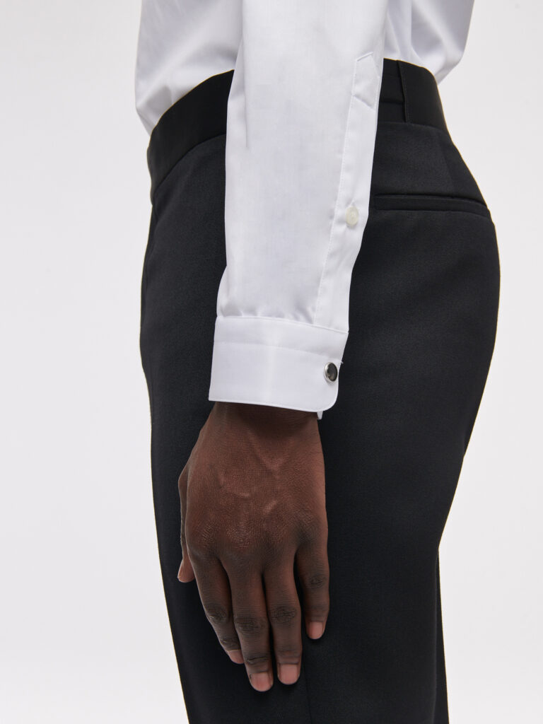 Manchette mousquetaire d'une chemise blanche fermée par un bouton de manchette noir, bras le long d'un pantalon noir