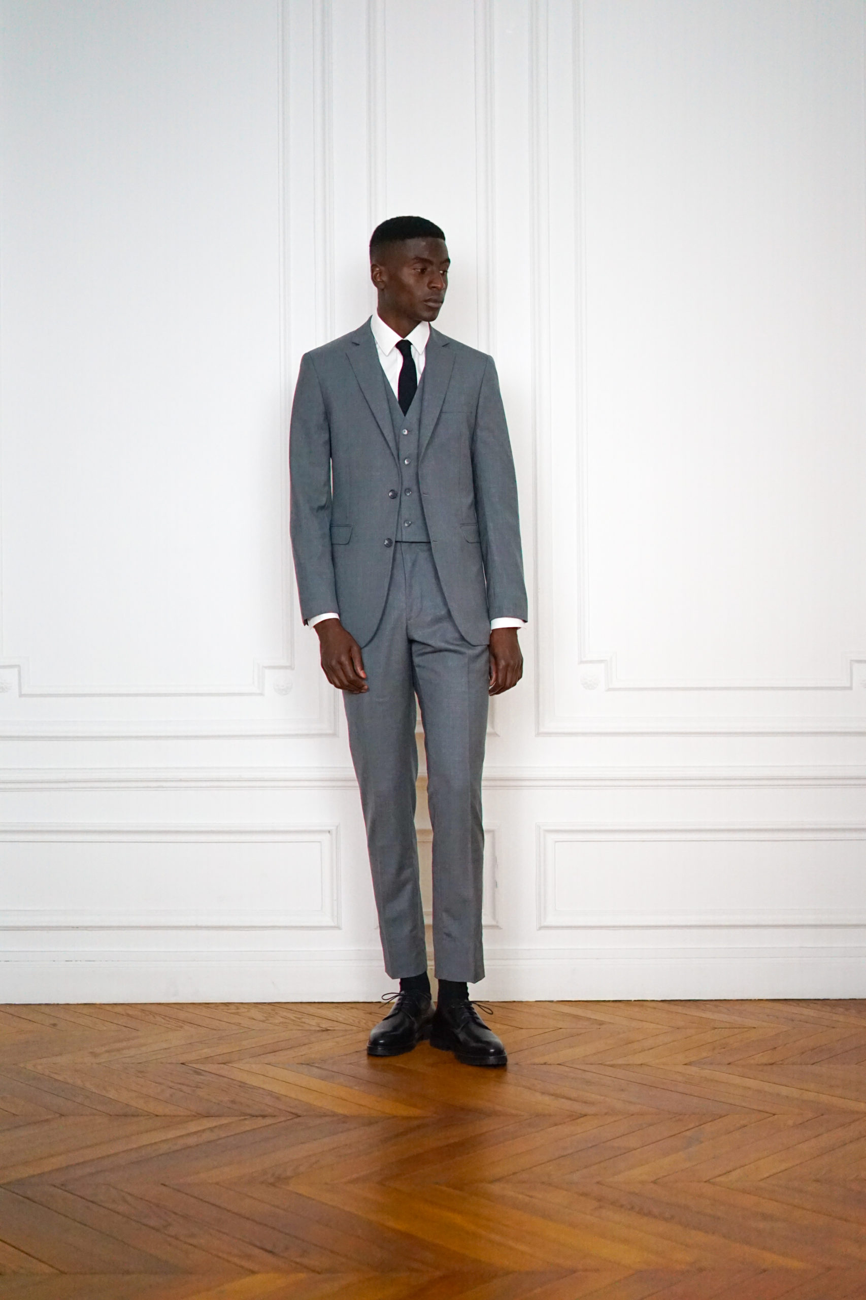 Costume trois pièces gris sur-mesure porté avec une chemise blanche et une cravate sombre, en pied devant des boiseries blanches, RIVES Paris