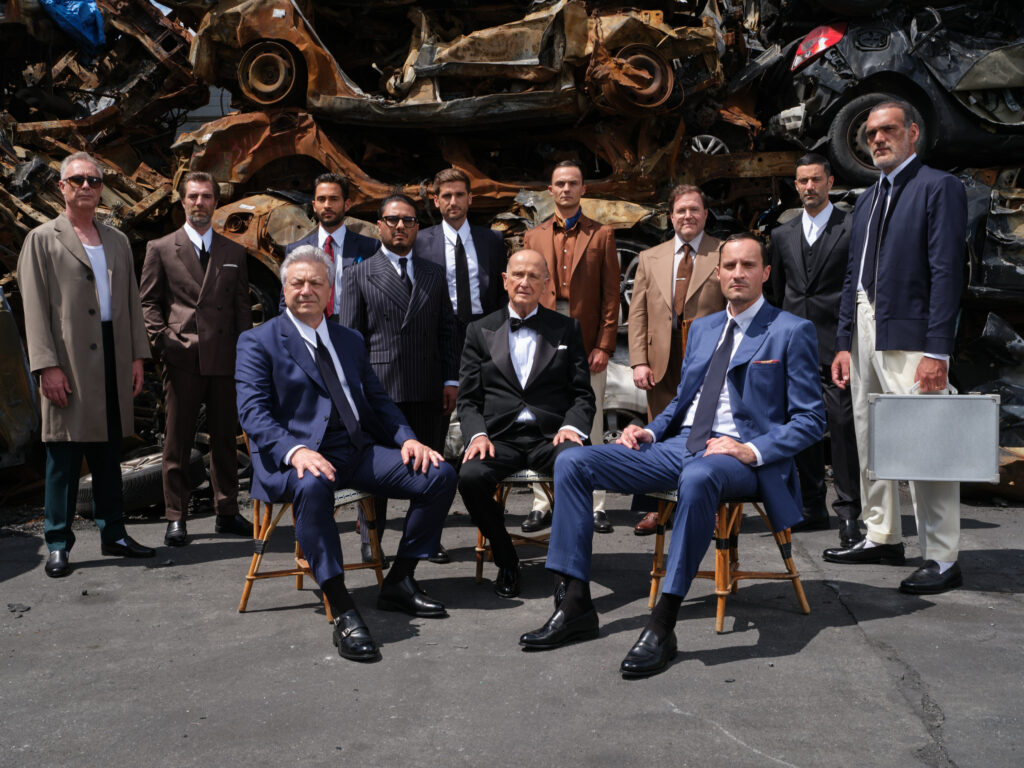 Groupe d'hommes en costumes sur-mesure RIVES Paris, devant un fond de voitures en métal, style élégant et contemporain.